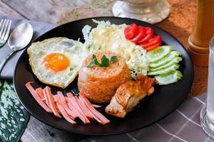 gebraten Reis, gebraten Ei, Gurke, geschnitten Tomate, Grün Salat und gebraten Hähnchen platziert auf ein schwarz Teller wie ein Garnierung. foto