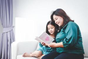 Mutter und Tochter sitzen im das Leben Zimmer auf das Sofa öffnen und lesen das Gruß Karte Das das wenig Mädchen schrieb zum Mama wie ein Geschenk. Familie Konzept Single Mutter foto