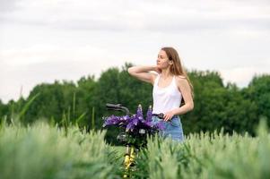 ein süß Mädchen ist Stehen mit ein Strauß von Lupinen im ein Feld in der Nähe von ein Fahrrad foto