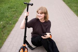 ein Teenager Mädchen ist Sitzung auf ein Roller mit ein Telefon und lächelnd foto