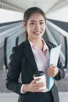 junge asiatische geschäftsfrau im schwarzen anzug, der ordner und tasse kaffee hält und auf gehwegstation steht. foto