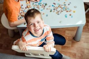 ein Junge von 6 Jahre alt sammelt ein Puzzle beim Zuhause und sieht aus beim das Kamera mit ein Lächeln foto