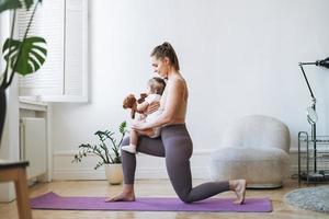 junge frau passte mutter mit babymädchen in ihren armen, die zu hause fitness auf der matte machen foto