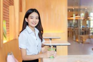 Porträt von süß ein asiatisch thailändisch Mädchen Schüler im Uniform ist Sitzung lächelnd glücklich und selbstbewusst erfolgreich während halten Glas von trinken im Kaffee Geschäft im Universität. foto