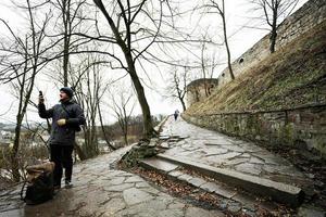 Mann Tourist mit Rucksack aussehen beim Telefon, Stand auf nass Pfad zu ein uralt mittelalterlich Schloss Festung im Regen. foto