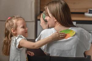 heiter wenig Mädchen geklebt ein Stück von Papier Fisch zu ihr Mutter auf ihr zurück beim Zuhause im das Küche. komisch Familie Witze, April Narren Tag Feier foto