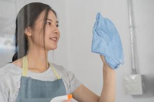 Reinigung Hygiene, lästig von asiatisch jung Maid Frau, Mädchen Hand im mit ein Wasser Sprühgerät Flasche zu wischen sauber, verwenden Blau Lappen abwischen beflecken auf Glas, Fenster im Restaurant. Hauswirtschaft sauber hoch, Reiniger foto