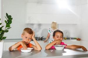 glücklich jung Familie, Mutter mit zwei Kinder, bezaubernd Kleinkind Mädchen und komisch unordentlich Junge haben gesund Frühstück Essen Obst und Molkerei, Sitzung im ein Weiß sonnig Küche mit Fenster foto