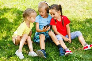 Unternehmen von drei Kinder im das Park sind Sitzung auf das Gras mit ein Telefon foto