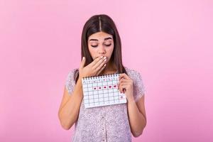 Porträt von ein schön jung Mädchen halten ihr Menstruation Kalender und Abdeckung hören Gesicht isoliert Über Rosa Hintergrund. Zeitraum Kalender foto