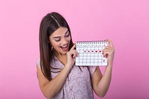Porträt von lächelnd jung Frau halten Zeitraum Kalender isoliert Über Rosa Hintergrund im Studio. schön jung Frau Stehen isoliert Über Rosa Hintergrund, zeigen Menstruation- Kalender foto