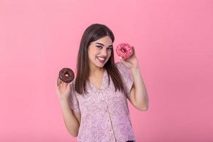 stilvoll Mädchen mit lange Haar positiv Posen, halten frisch Rosa und Schokolade Donuts mit Pulver bereit zu genießen Süßigkeiten. Porträt von attraktiv jung Frau im retro Hemd haben Spaß mit süße Sachen foto