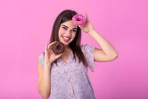 stilvoll Mädchen mit lange Haar positiv Posen, halten frisch Rosa und Schokolade Donuts mit Pulver bereit zu genießen Süßigkeiten. Porträt von attraktiv jung Frau im retro Hemd haben Spaß mit süße Sachen foto