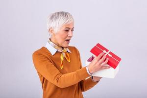 glücklich reifen Frau mit ein Geschenk. isoliert Über Hintergrund. schön stilvoll Senior Dame Öffnung ihr Weihnachten Geschenk. elegant reifen Frau halten ein rot Geschenk Box foto