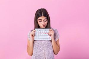 Porträt von enttäuscht empört Frau 20er Jahre halten Zeitraum Kalender isoliert Über Rosa Hintergrund im Studio. schön jung Frau Stehen isoliert Über Rosa Hintergrund, zeigen Menstruation- Kalender foto