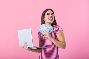 Porträt von ein aufgeregt zufrieden Mädchen halten Geld Banknoten und Laptop Computer. glücklich Frau mit Glücklich Geld. Foto von schön Frau mit Laptop, isoliert auf Rosa Hintergrund