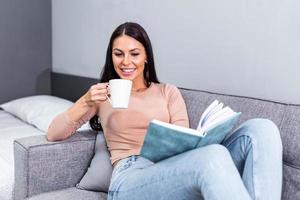 jung Frau beim Zuhause Sitzung auf modern Sofa entspannend im ihr Leben Zimmer lesen Buch und Trinken Kaffee oder Tee. gemütlich Bett und ein schön Mädchen, lesen ein Buch, Konzepte von Zuhause und Komfort foto