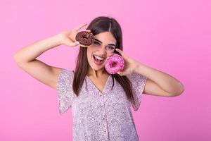 stilvoll Mädchen mit lange Haar positiv Posen, halten frisch Rosa und Schokolade Donuts mit Pulver bereit zu genießen Süßigkeiten. Porträt von attraktiv jung Frau im retro Hemd haben Spaß mit süße Sachen foto