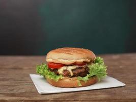groß Burger Lügen auf Kunst Weiß Papier gegen hölzern Tisch. ein saftig Grün Salat Blatt und ein rot Tomate Lüge in der Nähe von das Burger foto