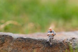 Miniaturgeschäftsmann, der eine Zeitung auf einem Felsen mit einem Naturhintergrund liest foto