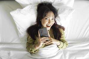 ein lächelnd jung asiatisch Frau ist Lachen während halten ihr Telefon und Lügen auf das Bett foto