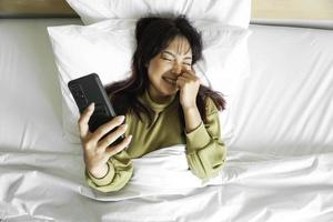 ein lächelnd jung asiatisch Frau ist Lachen während halten ihr Telefon und Lügen auf das Bett foto