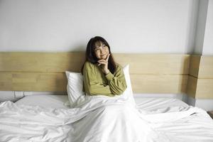 ein nachdenklich jung asiatisch Frau ist suchen beiseite und halten ihr Kinn während Sitzung auf das Bett foto