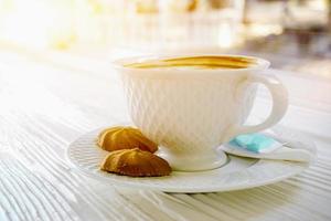 heiß Kaffee im ein Weiß Kaffee Tasse mit Kekse auf Weiß hölzern Tabelle auf Sonne Fackel Hintergrund. foto