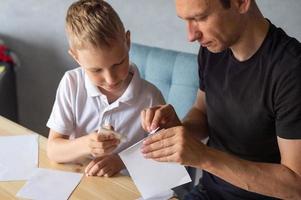 ein süß Junge ist Sitzung mit seine Papa beim das Tabelle und Sammeln Origami. hält ein Weiß Blatt von Papier foto