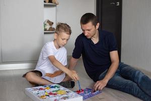 ein süß Junge im ein Weiß T-Shirt sammelt ein elektrisch Designer mit seine Papa im das Zimmer foto