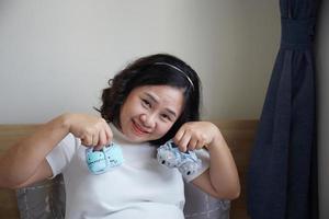 jung glücklich asiatisch schwanger halten klein Schuhe zum das ungeboren Baby im ihr Hand und aussehen in das Kamera foto