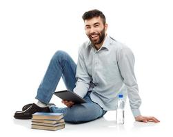 jung bärtig lächelnd Mann halten ein Tablette mit Bücher und ein Flasche von Wasser Sitzung auf ein Weiß foto