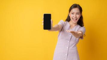 asiatische Frau, die auf ein leeres schwarzes Handy auf gelbem Hintergrund gestikuliert
