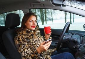 Geschäftsfrau im ein Pelz Mantel mit rot Lippen Senden ein Text Botschaft und Trinken Kaffee während Fahren foto