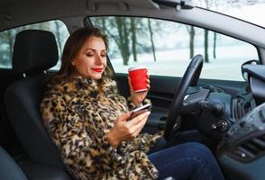 Geschäftsfrau im ein Pelz Mantel mit rot Lippen Senden ein Text Botschaft und Trinken Kaffee während Fahren foto