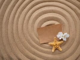 Brief im das Sand. Kunst Papier mit ein Weiß Blume und ein Seestern auf Sand im das gestalten von ein Spiral. Strand Urlaub, Zen foto