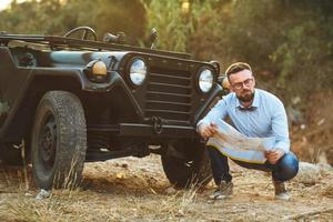 jung stilvoll Mann mit Brille und Bogen Krawatte sehen das Karte in der Nähe von das altmodisch suv foto