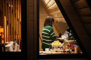 Frau bereitet vor Abendessen im das Küche, ein Aussicht durch das Fenster von ein Land Haus beim Nacht. foto