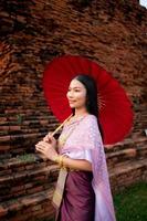 schön thailändisch Mädchen im traditionell Kleid Kostüm rot Regenschirm wie thailändisch Tempel wo ist das Öffentlichkeit Ort, thailändisch Frau im traditionell Kostüm von Thailand. foto