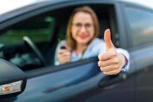 jung glücklich Frau Sitzung im ein Auto mit das Schlüssel im ihr Hand und Daumen oben foto