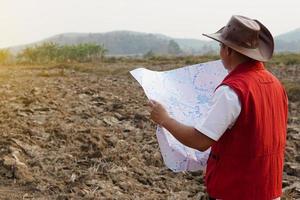 asiatisch Mann Forscher trägt Hut, rot Weste Shirt, hält Karte zu erkunden Land Grenze. Konzept, Land Planung, erkunden Eigentum. geodätisch Umfrage Bereich. Schatz Jagd auf Land. Abenteuer Lebensstil. foto