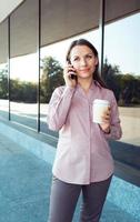 Geschäftsfrau mit Handy und Kaffee während Stehen gegen Büro Gebäude, draussen foto