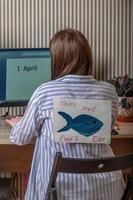 ein jung Freiberufler sitzt mit ihr zurück zu uns beim ein Computer, ein Papier Fisch ist befestigt zu ihr zurück. April Narren Tag, Streich jeder andere foto