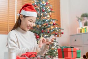 feiern auf Weihnachten Vorabend, Neu Jahr Jahreszeit, süß asiatisch jung Frau, Mädchen halten Smartphone nehmen Foto rot Geschenk Kasten, erhalten Geschenk auf fröhlich Weihnachten Tag. Glücklich, heiter auf Festival von Menschen, Technologie.