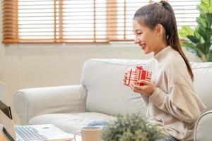 feiern auf Tag von Liebe, Neu Jahr Tag Jahreszeit, ziemlich asiatisch jung Frau, Mädchen mit Laptop Computer, Gruß auf Video Anruf während Sozial Distanz, halten Geschenk Kasten, glücklich zu erhalten Geschenk auf Valentinstag. foto