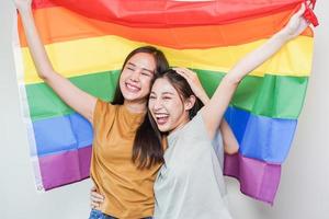 glücklich lesbisch, schön asiatisch jung zwei Frauen, Mädchen Fröhlich, Paar Liebe Moment Ausgaben gut Zeit zusammen, halten oder winken lgbt Regenbogen, Stolz Flagge auf isoliert Weiß Mauer Hintergrund zusammen beim heim. foto