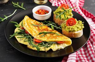 Frühstück. Omelette mit Tomaten, Käse, Grün Rucola und Toast mit Avocado Sahne auf schwarz Platte. Frittata - - Italienisch Omelett. foto