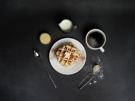oben Aussicht von lecker Waffeln Platte, Karamell Soße, Kaffee Tasse, Milch, Dessertlöffel, Sieb auf ein schwarz Beton Hintergrund foto