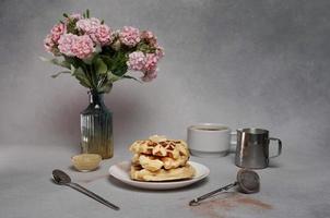 hausgemacht von lecker Waffeln Platte, Karamell Soße, Kaffee Tasse, Milch, Rosa Blumen auf ein grau Hintergrund foto