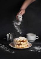 kaukasisch Mädchen Hand Sträusel Zucker Pulver auf köstlich Waffeln, Karamell Soße, Kaffee Tasse, Milch, Dessert Löffel, Sieb auf schwarz Beton Hintergrund foto